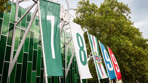 Mehrere große Banner mit den Zahlen 7, 8, 9 und 0 hängen an einem Zaun vor Bäumen