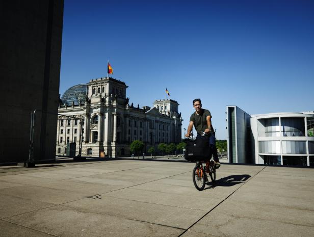 Der Autor Hasnain Kazim fährt mit einem Fahrrad auf einem Betonplatz vor dem Reichstagsgebäude in Berlin