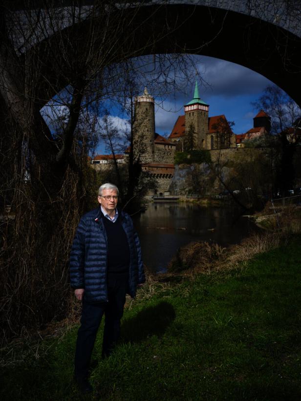 Ein Mann steht im Anorak unter einer Steinbrücke, im Hintergrund ist eine Kirche zu sehen