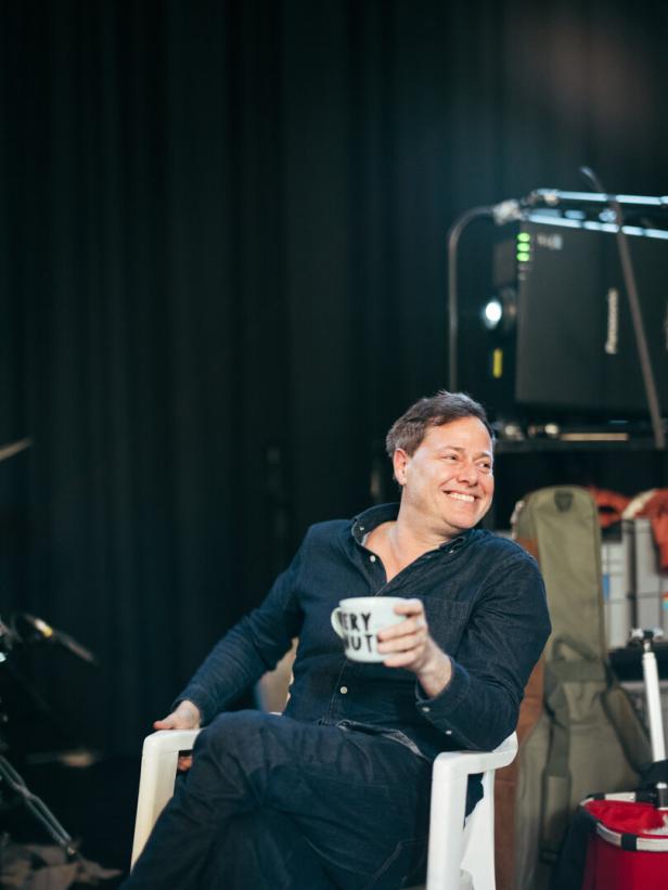 Ein lachender Milo Rau mit Kaffeetasse in der Hand, in einem dunklen Theaterprobenraum 
