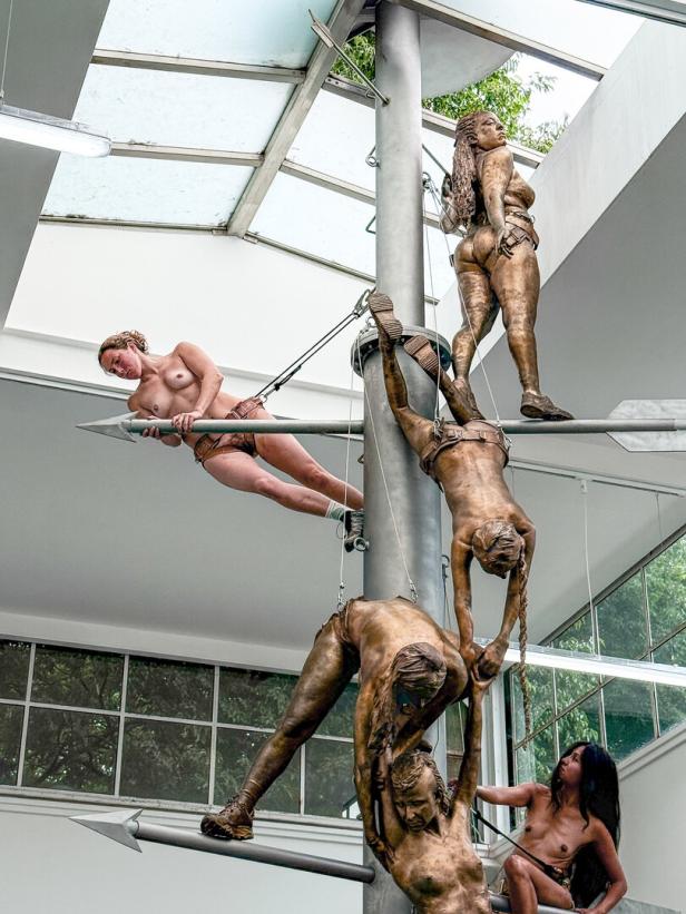 Eine Reihe unbekleideter Performerinnen klettert in einem Innenraum eine mit Skulpturen dekorierte Säule hinauf.