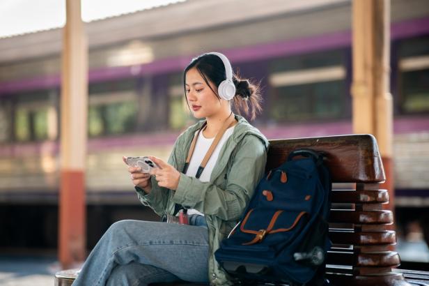Frau mit Kopfhörern sitzt auf einer Bank am Bahnsteig und schaut auf ihr Smartphone 
