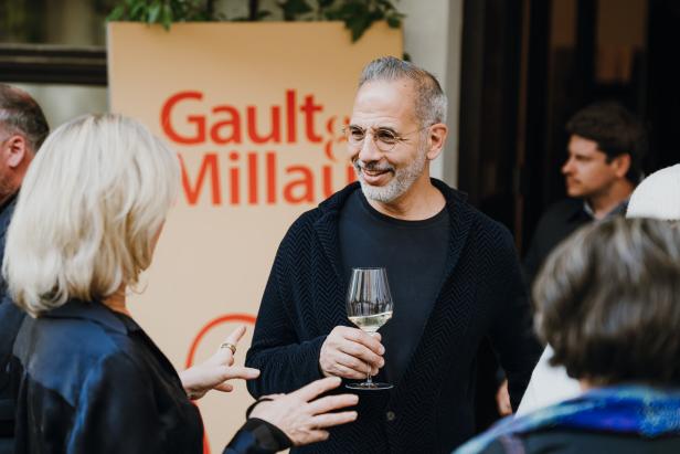 Yotam Ottolenghi steht mit einem Glas in der Hand vor einem Gault-Millau-Plakat und unterhält sich mit einer blonden Frau