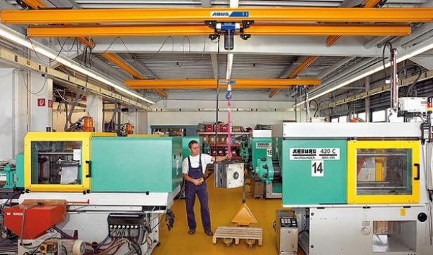 Mann in Arbeitskleidung bedient Hebekran über zwei großen Spritzgießmaschinen in einer hellen Fabrikhalle.