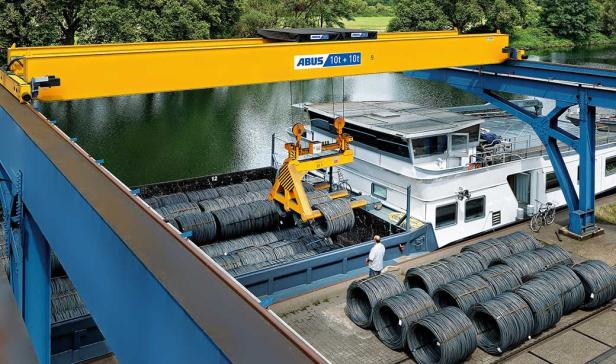Gelber Laufkran mit der Aufschrift 'ABUS 10t + 10t' hebt Stahlrollen über einem Schiff im Hafen.