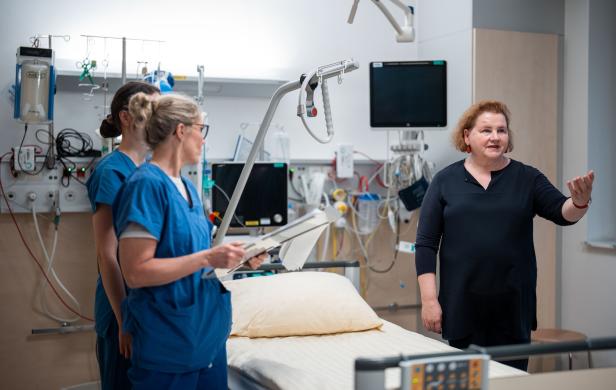 In einem Krankenzimmer erklärt eine Frau in Schwarz zwei Krankenschwestern in blauer Arbeitskleidung etwas. Es ist Sozialministerin Schumann..
