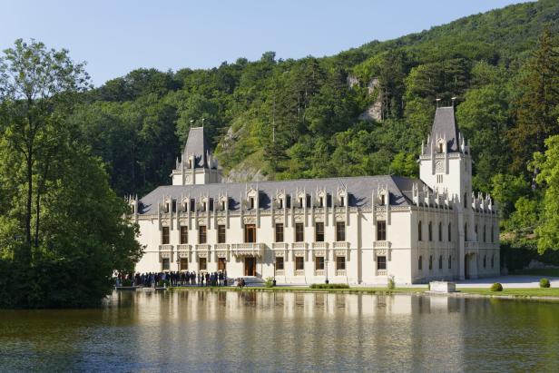 Ein großes, helles Schloss mit Türmen steht vor einem Teich, umgeben von dichtem grünen Wald.