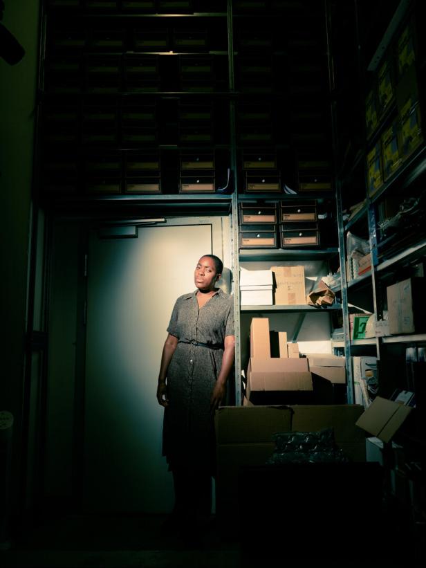 Die Künstlerin Onyeka Igwe in einem halbdunklen Raum, in den Regalen Bücher, Kisten, Akten