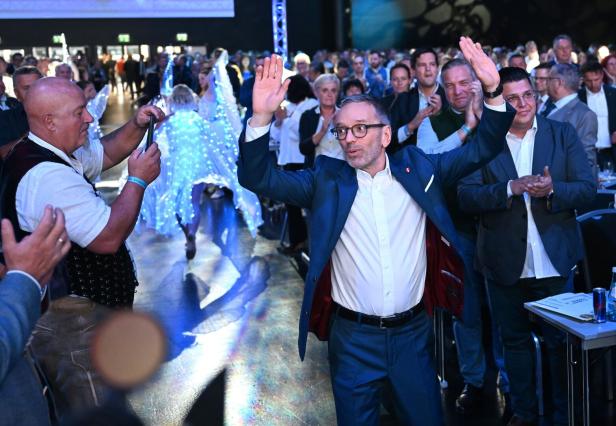 Herbert Kickl winkt in einem blauen Anzug vor einem applaudierenden Publikum, während links eine Person mit dem Smartphone filmt.