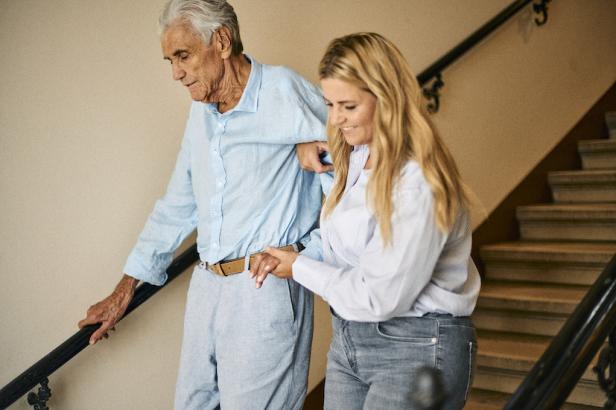 junge blonde Frau hilft älterem weißhaarigen Mann die Treppen hinunter
