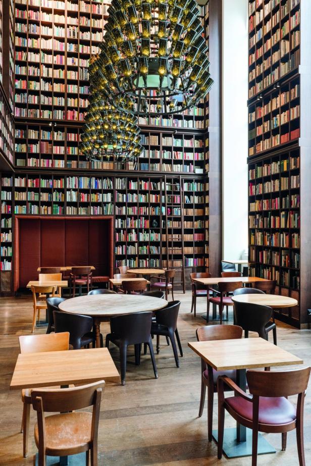 Großer, hoher Lesesaal mit bodenhohen Bücherregalen, runden und quadratischen Holztischen sowie einem großen, modernen Kronleuchter
