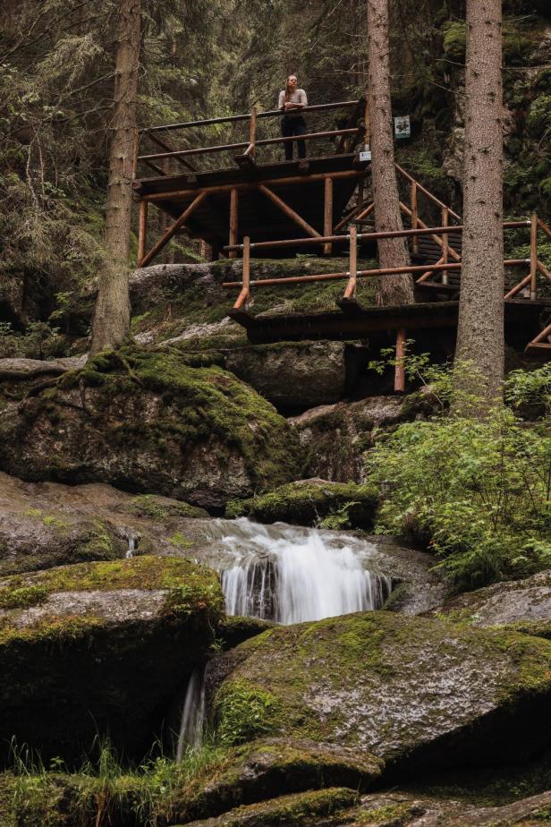 Person steht auf einer hölzernen Aussichtsplattform im Wald über einem kleinen Wasserfall und moosbedeckten Felsen