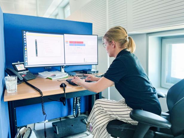 Frau mit Brille arbeitet an zwei Monitoren an einem Bürotisch mit Telefon und Tastatur.