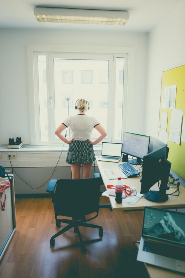 Eine Frau steht in einem Büro am Fenster, vor ihr Schreibtisch mit Monitoren, Laptop und Stuhl.