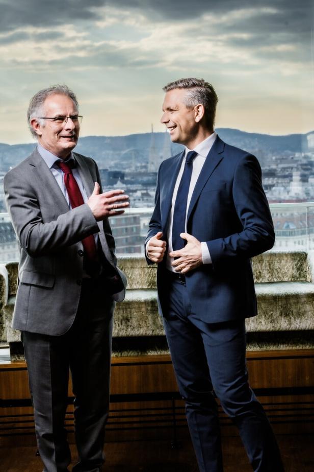 Zwei Männer in Anzügen, die Minister Marterbauer und Hattmannsdorfer,  unterhalten sich vor einem Fenster mit Blick auf eine Stadt.