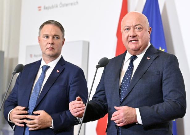 Zwei Männer in Anzügen, Vizekanzler Babler und Bundeskanzler Stocker, stehen vor Mikrofonen und österreichischen sowie europäischen Flaggen.