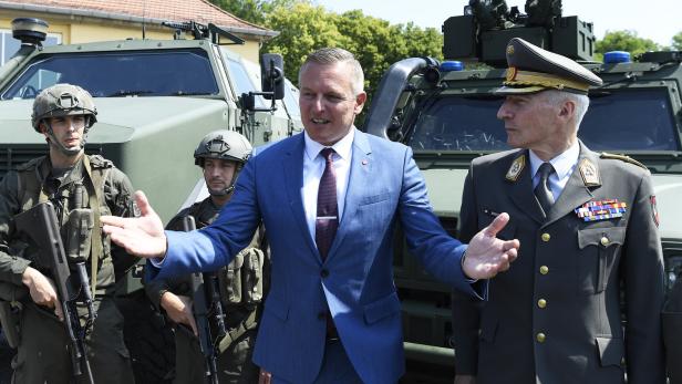 Mario Kunasek und ein Offizier stehen vor Militärfahrzeugen, flankiert von Soldaten.