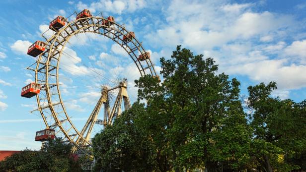 Das Wiener Riesenrad vor blauem Himmel mit Wolken, teilweise von Bäumen verdeckt.