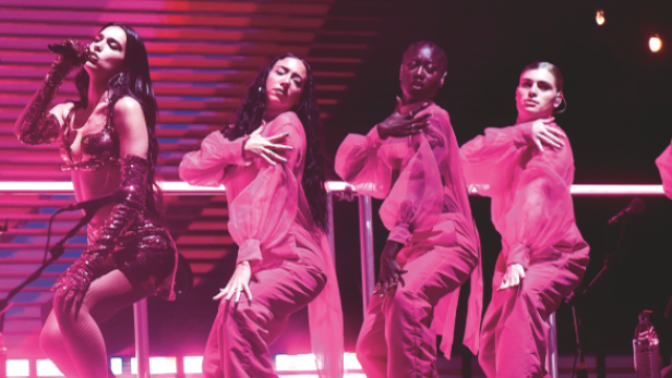 Dua Lipa tritt mit Tänzern in pinkfarbenen Overalls auf einer Bühne auf.