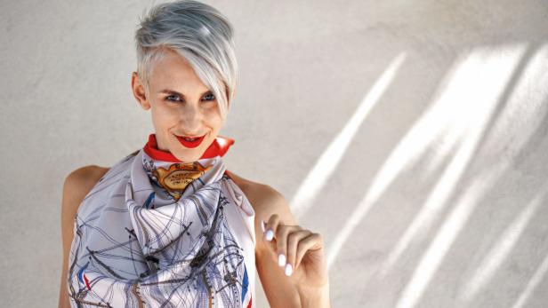 Eine Frau mit grauen Haaren und rotem Lippenstift trägt ein Tuch mit Schiffsdesign.