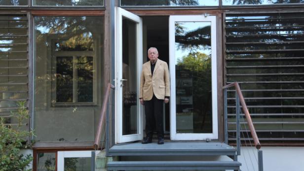 Ein älterer Mann steht auf der Treppe vor einem modernen Haus mit vielen Fenstern.