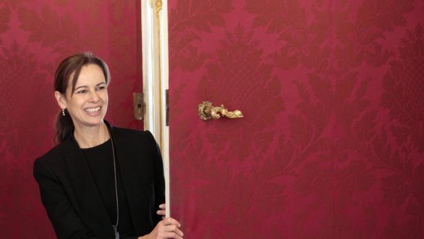 Eine lächelnde Frau in einem schwarzen Blazer steht in einer Türöffnung vor einer roten Wand.