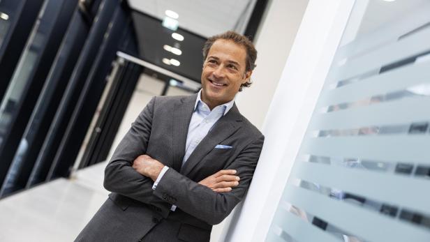 Ein lächelnder Mann im Anzug lehnt an einer Wand in einem Bürogebäude.