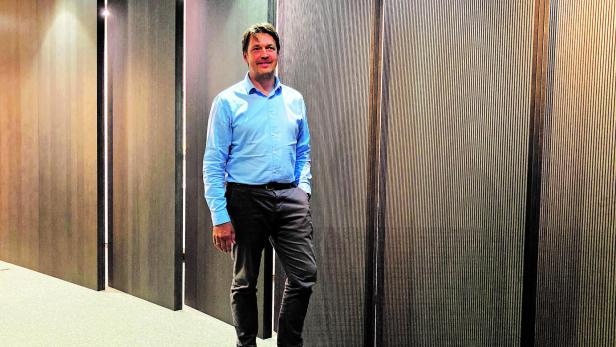 Ein Mann in hellblauem Hemd posiert vor einer Wand mit vertikalen Linien.