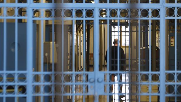 Eine Person steht hinter blauen Gitterstäben in einem hellen Flur mit mehreren Türen und Fenstern.