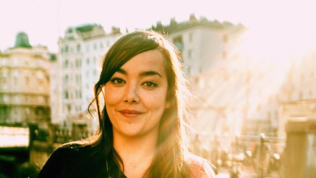 Eine junge Frau lächelt vor einer Stadtkulisse im goldenen Abendlicht.