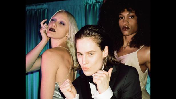 Christine and the Queens posiert mit zwei weiteren Personen vor einem Vorhang.