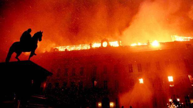 Ein Gebäude steht in Flammen, im Vordergrund eine Reiterstatue im Gegenlicht.
