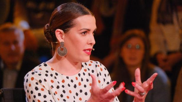 Eine Frau mit Ohrringen und gepunktetem Oberteil gestikuliert während einer Präsentation.