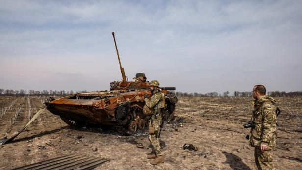 Zwei Soldaten stehen vor einem zerstörten Panzer auf einem Feld.