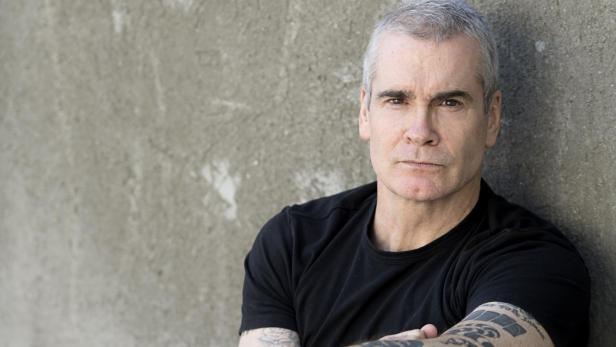 Porträt von Henry Rollins, der an einer Wand sitzt.