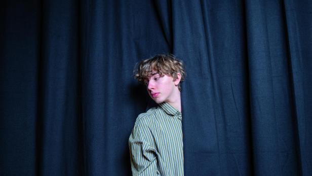 Ein junger Mann mit gestreiftem Hemd steht hinter einem dunklen Vorhang.
