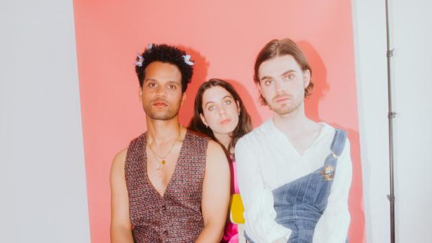 Drei Personen posieren vor einem rosa Hintergrund im Studio.