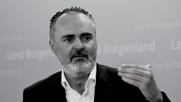 Ein Mann mit Bart gestikuliert vor einem Hintergrund mit der Aufschrift „Land Burgenland“.