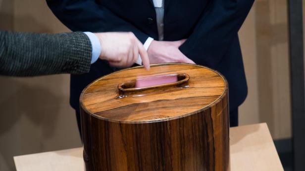 Eine Person wirft einen Stimmzettel in eine hölzerne Wahlurne.