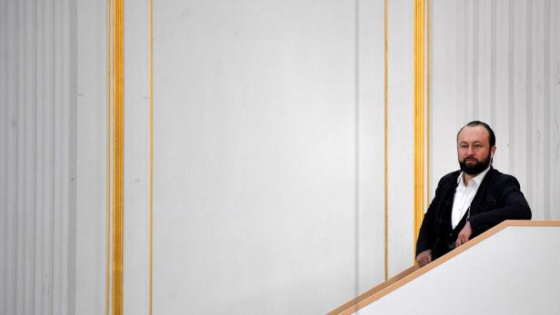 Ein Mann mit Bart lehnt an einem Treppengeländer vor einer verzierten Wand.