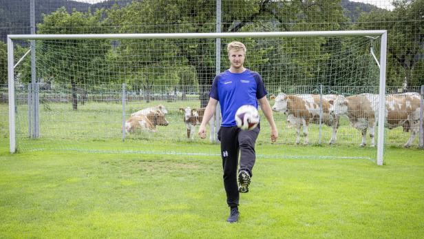 Ein Mann jongliert einen Fußball vor einem Fußballtor, hinter dem Kühe grasen.