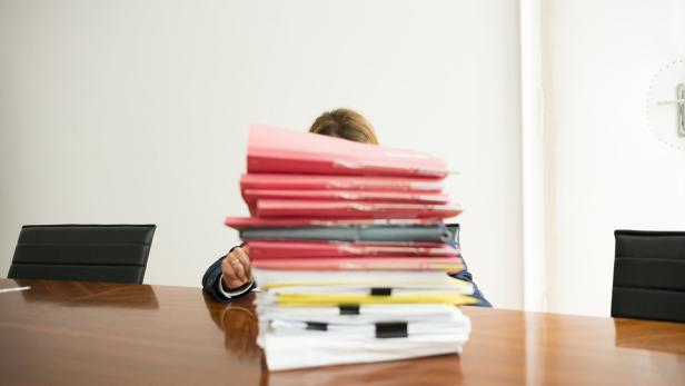 Eine Person sitzt hinter einem hohen Stapel Aktenordner an einem Schreibtisch.