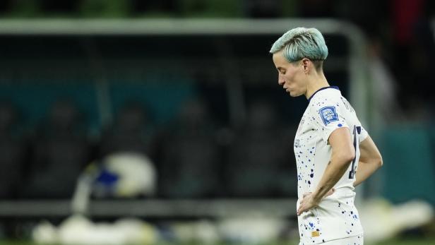 Megan Rapinoe mit blau gefärbten Haaren steht mit gesenktem Kopf auf dem Spielfeld.