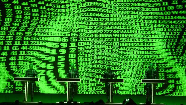 Die Band Kraftwerk tritt vor einer Leinwand mit grünen, binären Codes auf.