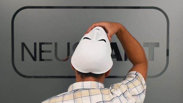 Eine Person hält eine weiße Maske vor dem Kopf, im Hintergrund ein Logo mit der Aufschrift „NEUART“.