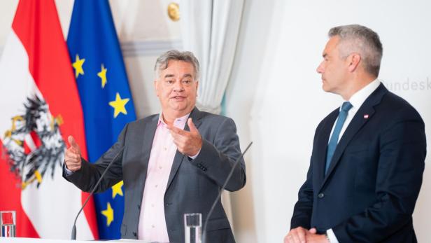 Zwei Männer stehen vor Flaggen des Bundeskanzleramts, Österreichs und der EU.