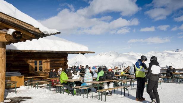 Skifahrer sitzen an Tischen vor einer Hütte in den Bergen.