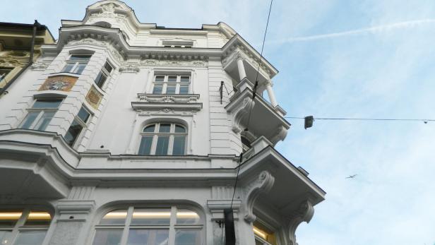 Ein weißes, reich verziertes Gebäude mit großen Fenstern und Balkonen vor blauem Himmel, im Hintergrund fliegt ein Flugzeug.