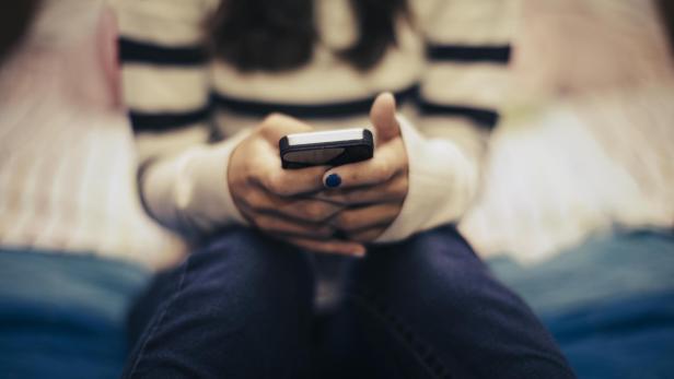 Eine junge Frau sitzt mit ihrem Smartphone in der Hand auf einem Bett.