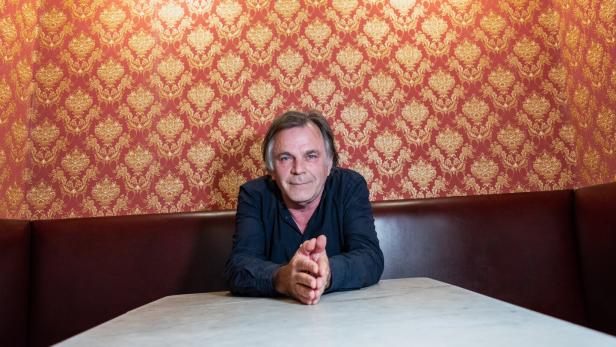 Markus Hinterhäuser sitzt an einem Tisch vor einer Wand mit auffällig orangem Muster.